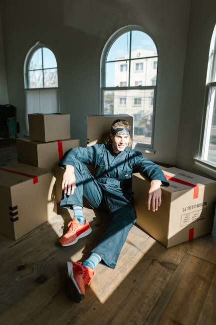 A person dressed in casual clothing, including a dark jacket, blue pants, and bright orange sneakers, is sitting on the floor inside a room with large arched windows that let in natural light. The ind