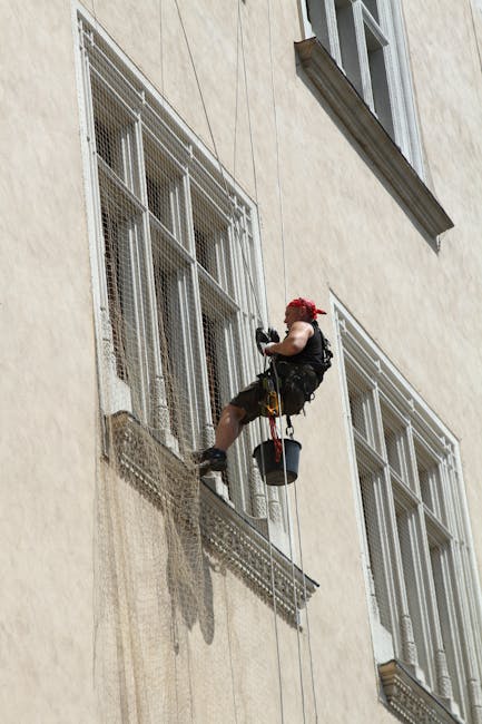 A professional window cleaner is suspended on a harness attached to a rope, cleaning the exterior of a multi-storey building with a scaffolding platform in Finsbury Park. The worker wears a red safety helmet, gloves, and work attire, holding cleaning tools in hand. The building's façade features large, barred windows with mesh screens and beige stone walls. The window cleaning equipment includes a bucket hanging from the harness, positioned near the worker's waist, and cleaning cloths or brushes in hand. Sunlight illuminates the scene, casting shadows on the building's exterior, while the worker's activity aligns with ongoing building maintenance or interior access preparation commonly associated with house removals and property management. Finsbury Park's professional cleaning services often support property preparation during home relocation or moving processes, as indicated by the clean, accessible windows visible in the image.
